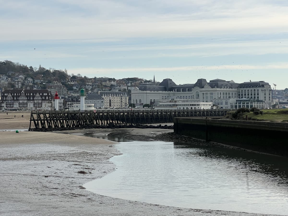 Chenal à marée basse entre des bancs de sable, une jetée de bois et deux petits phares, la ville de Trouville en arrière-plan.