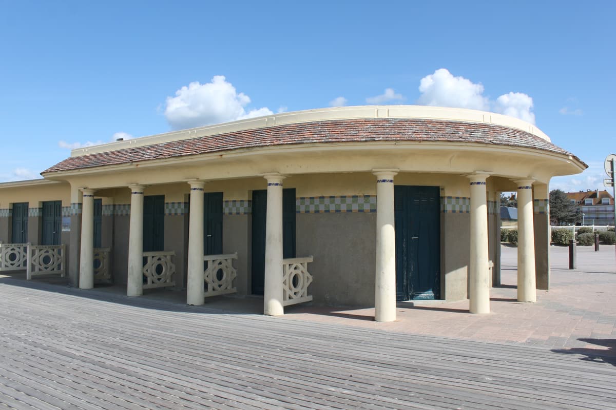 Pavillon arrondi à colonnes et portes vertes, couvert de tuiles, au bord de la promenade en planches, sous un ciel bleu.