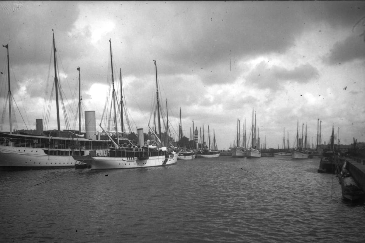 Photographie ancienne : des yachts à vapeur et des voiliers à quai dans un bassin, sous un ciel nuageux.