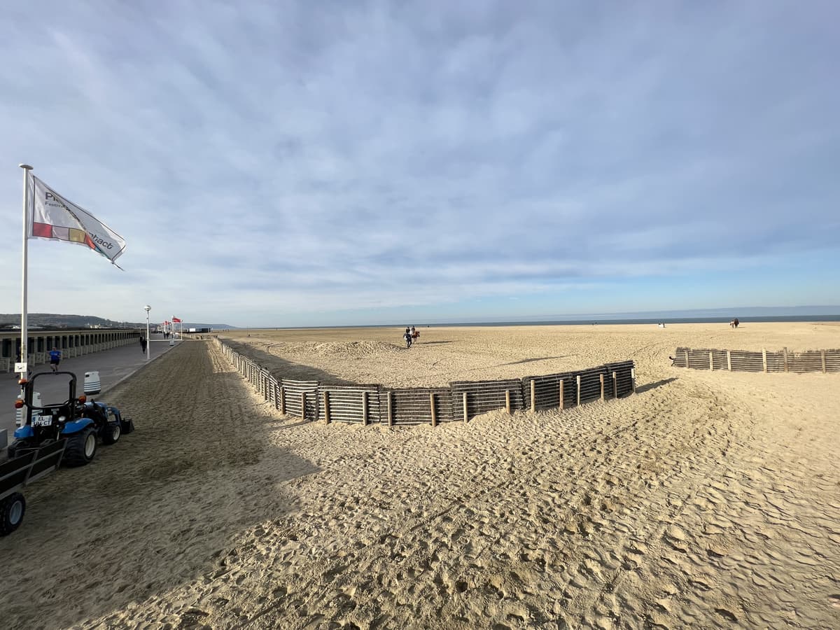 Grande plage de sable presque vide, une palissade de ganivelles, un drapeau et la ligne des cabines à gauche.