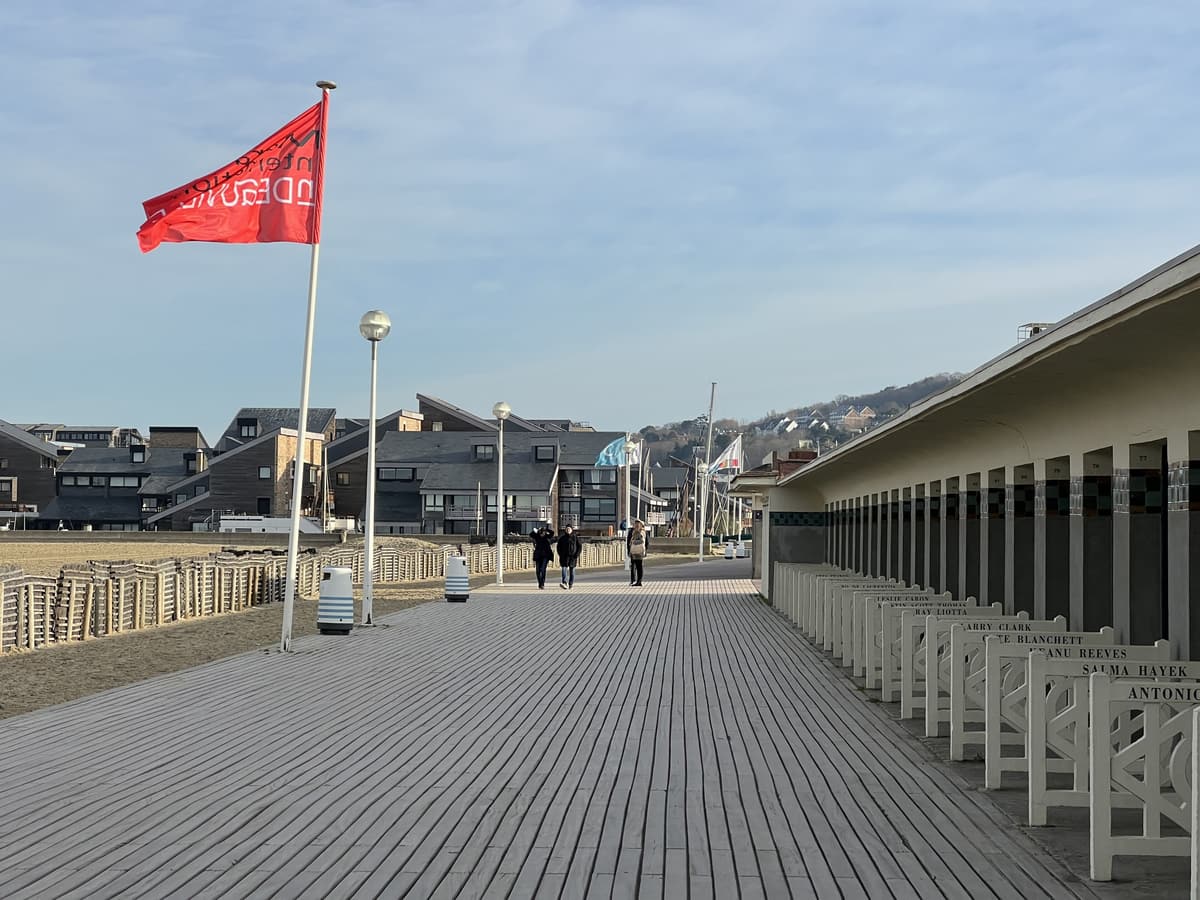 Longue promenade en planches de bois le long d'une rangée de cabines blanches, un drapeau rouge, la plage à gauche et des immeubles au loin.