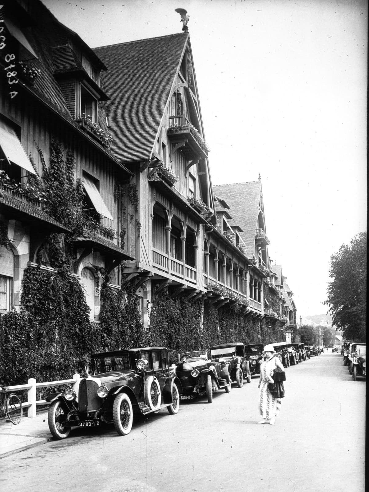 Photographie ancienne en noir et blanc : une rue bordée de grands immeubles à pans de bois et balcons fleuris, des automobiles des années 1920 garées le long du trottoir.