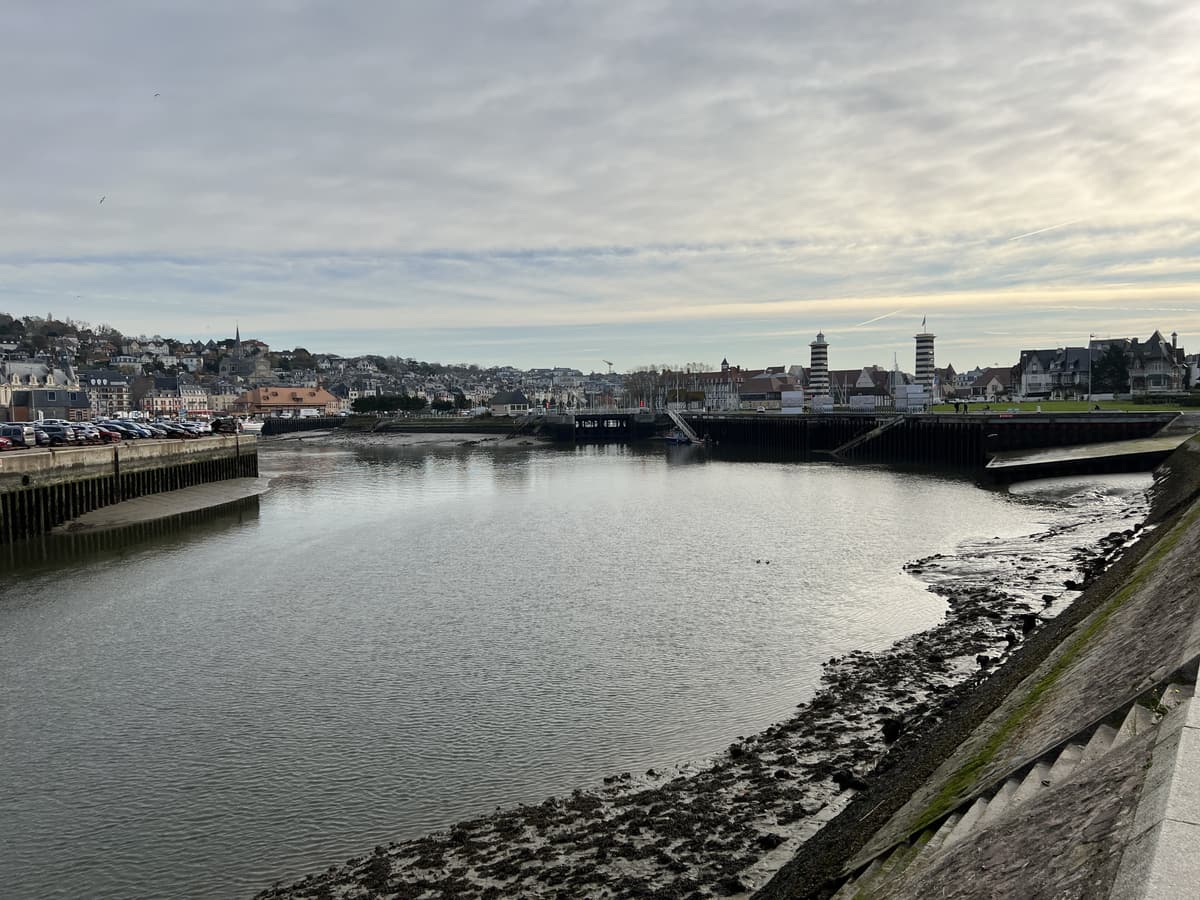 La Touques à marée basse entre un quai et une berge de rochers, avec la ville en arrière-plan sous un ciel gris.